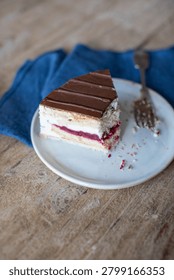 High-angle view of a single slice of layered mousse cake or gateau served on a small ceramic plate. The dessert features distinct layers of sponge cake, white cream, and a dark berry or cherry filling, topped with a smooth layer of chocolate glaze and a fork on a rustic wooden table with a blue napkin.
