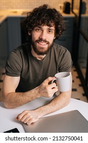 High-angle View Of Smiling Bearded Young Business Man Holding In Hand Cup With Morning Coffee Sitting At Table With Closed Laptop Computer, Looking At Camera, In Kitchen With Modern Interior.