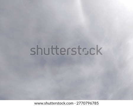 A high-angle view of a soft, hazy sky filled with delicate, wispy cirrus and cirrostratus clouds. 

The color palette is a gentle blend of white and various shades of pale grey, creating a serene and ethereal atmosphere. In the upper right quadrant, a faint but distinct vertical band of spectral colors, known as a sun dog or parhelion, is visible. 

This rare optical phenomenon occurs when sunlight refracts through ice crystals suspended high in the atmosphere, casting a subtle rainbow-like shimmer against the clouds.

The overall image captures a moment of quiet, natural beauty in the heavens, suitable for use as a background or to illustrate atmospheric and meteorological themes.