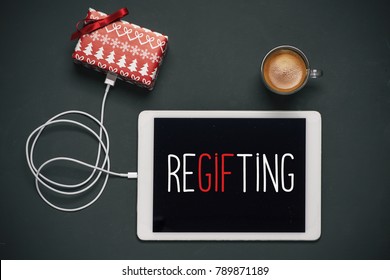 High-angle View Of A Tablet With The Word Regifting In Its Screen, Connected To A Gift By A Cable, And A Cup Of Coffee, Placed On A Dark Gray Table