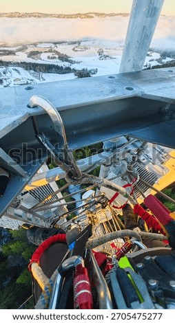 High-angle view from a telecom tower showing safety harness, carabiners and ladder, with snowy farmland and low clouds beyond.