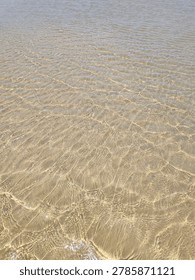 A high-angle view of a textured sandy beach during low tide, with a black buoy on a rope and a distant figure walking near a rocky reef.