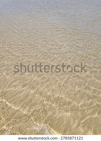 A high-angle view of a textured sandy beach during low tide, with a black buoy on a rope and a distant figure walking near a rocky reef.