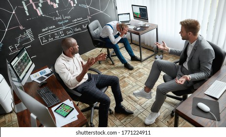 High-angle view of three traders sitting by desks in front of computer monitors while working in the office. Blackboard full of charts and data analyses in background. Stock trading, people concept.