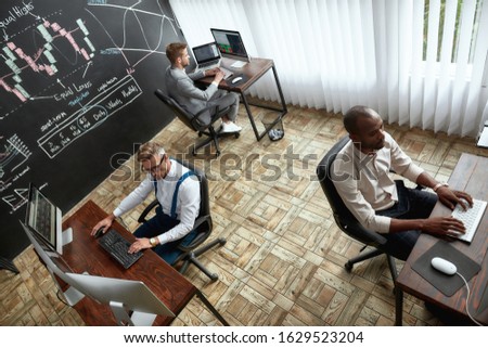 High-angle view of three traders sitting by desks in front of computer monitors while working in the office. Blackboard full of charts and data analyses in background. Stock trading, people concept.