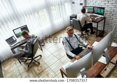 High-angle view of three traders sitting by desks in front of computer monitors while working in the office. Stock trading, people, business concept.