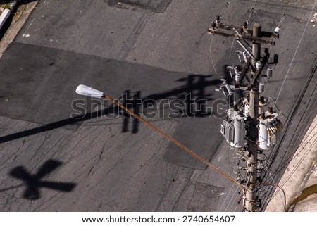 High-angle view of a utility pole with transformers and a street lamp, casting strong shadows on the asphalt. A concept of urban infrastructure, power distribution, and city textures.