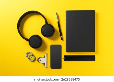 A high-contrast flat lay of school and tech essentials including black headphones, a notebook, smartphone, pen, ruler, clips, and binder arranged neatly on a bright yellow surface. 