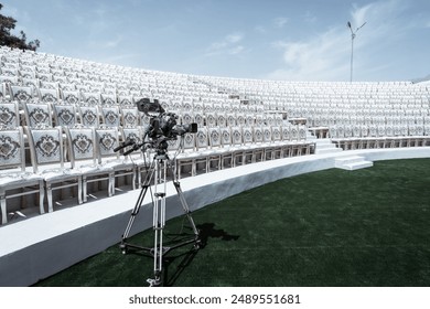 High-definition video camera on a tripod set up in an empty amphitheater with rows of ornate, white chairs. The scene features a clear blue sky, green artificial grass, and modern lighting fixtures - Powered by Shutterstock