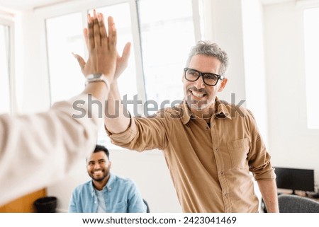 A high-five between colleagues, mature man in focus, emphasizes the exuberant success and positive reinforcement prevalent in their modern workspace. The high energy and team-centric atmosphere