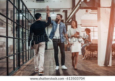 High-five!  Full length of two cheerful young business people giving high-five while their colleague looking at them and smiling