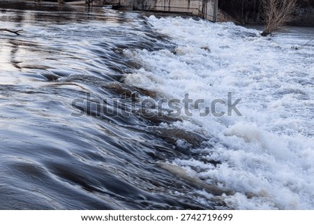 High-flow river waterfall during spring thaw, showing the transition from smooth laminar flow to intense turbulent whitewater after snowmelt. Powerful water surge cascading over a drop, with strong currents, swirling foam, and dynamic motion. Natural mountain river environment, dramatic hydrodynamic contrast, and high-energy water movement