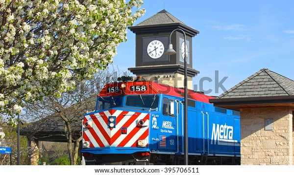 Highland\
Park, IL, USA - May 7, 2015: A springtime late afternoon view of a\
Metra commuter train leaving the Highland Park station, with a\
clock tower and spring white tree blossoms.\
\
