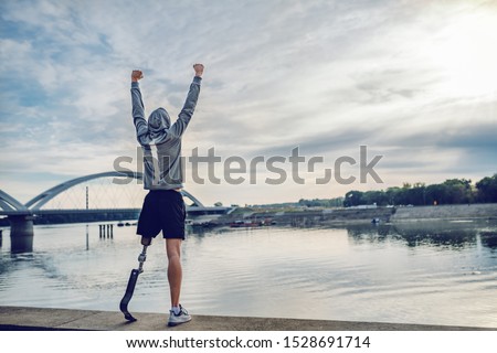 Highly motivated caucasian handicapped sportsman in sportswear and with artificial leg standing on quay and holding arms up. Only way to win is to be yourself.