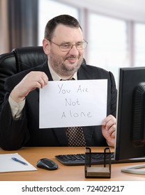 Highly Trained Male Psychiatrist With Pleasant Smile Shows Written Message You Are Not Alone To Desktop Monitor During Video Conference. 55 Years Old Bearded Man In Glasses Sits At Wooden Desk.