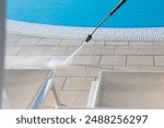 A high-pressure water jet cleans the tiles around a shimmering blue outdoor swimming pool, focusing on cleanliness and maintenance in a sunny, outdoor setting. Routine pool care and hygiene.