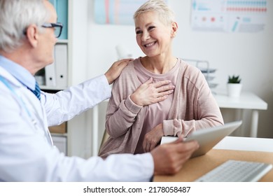 High-qualified Senior Doctor In Lab Coat Sitting At Table And Using Tablet While Reporting Good Tests Results To Senior Patient