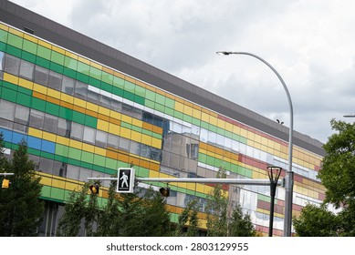 A high-quality, horizontally-oriented architectural photograph capturing the vibrant, modern facade of a contemporary building. The exterior wall is covered in an eye-catching, multi-colored grid pattern of glass and composite panels in shades of lime green, bright yellow, sky blue, and crimson red. Large bands of mirrored reflective windows run horizontally across the facade, mirroring the overcast sky and clouds overhead, as well as parts of neighboring concrete buildings. In the foreground, municipal infrastructure elements are visible, including a gray metal street light pole, a horizontal traffic arm with a pedestrian crossing signal icon, and lush green deciduous and evergreen tree branches framing the bottom and sides. This clean, geometric architectural composition is perfect for urban design portfolios, commercial construction brochures, civil engineering presentations, real estate mockups, and vibrant city-themed backgrounds.