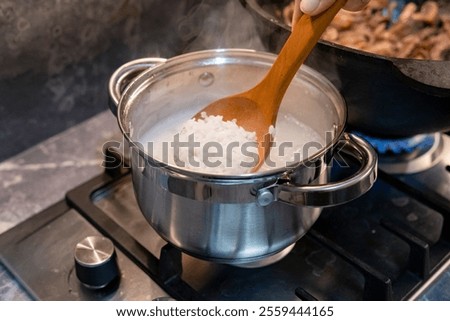  A high-quality, top-view shot capturing the process of cooking rice in an aluminum saucepan on a gas stove. The image shows grains of white rice simmering in boiling water, representing a fundamental step in preparing Asian cuisine, traditional risotto, or a healthy side dish. This authentic 