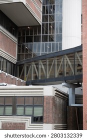 A high-quality, vertically-oriented architectural photograph showcasing a complex, multi-layered view of a modern institutional or university building complex. The central focus is an enclosed glass pedestrian skywalk or pedway bridge supported by white, repeating V-shaped structural steel trusses, spanning between a brutalist-style red brick facility and a cylindrical white concrete tower structure. Through the reflective glass panels of the skybridge, the dark silhouette of a single pedestrian can be seen walking across. The composition features a striking mix of building textures, including multi-paned glass curtain walls, red brick masonry, fluted concrete accents, and modern window frames. This geometric, urban architecture image is highly suitable for commercial use in engineering brochures, real estate marketing, construction company profiles, institutional design assets, and academic or medical campus conceptual layouts.