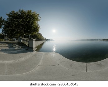 High-resolution 360-degree panoramic view of a tranquil waterfront promenade featuring elegant stone steps, decorative balustrades, tree-lined walkways, and calm reflective water under a clear blue sky. The scenic lakeside setting combines classical architecture with natural beauty, creating an inviting atmosphere for relaxation, tourism, recreation, and urban landscape photography. Suitable as an HDRI environment map, virtual reality background, or architectural visualization resource.