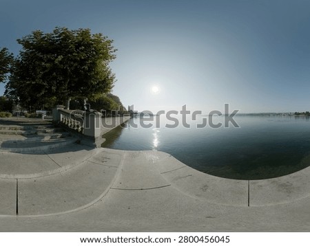 High-resolution 360-degree panoramic view of a tranquil waterfront promenade featuring elegant stone steps, decorative balustrades, tree-lined walkways, and calm reflective water under a clear blue sky. The scenic lakeside setting combines classical architecture with natural beauty, creating an inviting atmosphere for relaxation, tourism, recreation, and urban landscape photography. Suitable as an HDRI environment map, virtual reality background, or architectural visualization resource.