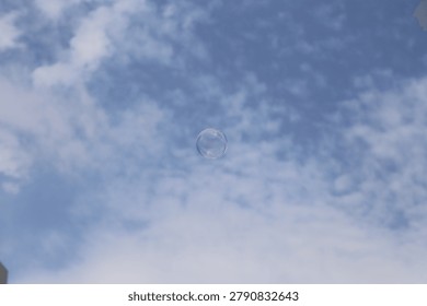 A high-resolution, eye-level shot capturing a single, iridescent soap bubble floating solitary in the center of the frame against a backdrop of a bright blue sky with scattered wispy white clouds. The mood is peaceful, whimsical, and ethereal, evoking themes of fragility, childhood, and fleeting moments. This image is ideal for advertising campaigns related to joy, purity, wellness, clean products, or summer lifestyle content.