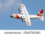 A high-resolution image of a C-130 Hercules aircraft in flight against a clear blue sky, showcasing its propeller engines, bold markings, and powerful design. 