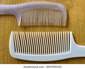 A high-resolution, overhead photograph capturing two plastic combs placed parallel on a warm, textured wooden surface. The top comb is translucent, frosted lavender-pink, while the bottom comb is opaque white