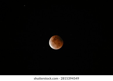 A high-resolution photograph of a partial lunar eclipse featuring a reddish moon illuminated against a dark starry night sky. The image captures the dramatic transition of Earth's shadow across the lunar surface, revealing detailed textures and natural colors. Perfect for astronomy, space science, celestial events, educational materials, lunar calendars, news articles, backgrounds, and creative designs with generous copy space.