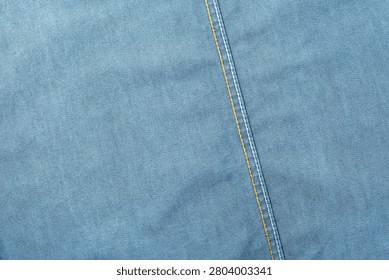 High-resolution texture background of smooth light wash blue denim. Features a prominent slanted vertical seam detailed with parallel lines of contrast yellow and white stitching.