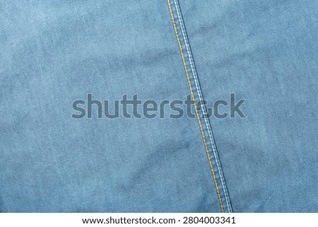 High-resolution texture background of smooth light wash blue denim. Features a prominent slanted vertical seam detailed with parallel lines of contrast yellow and white stitching.