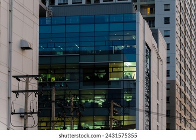 A high-rise building with a glass facade and a power pole in the foreground. A businessperson is standing in an office. The building is located in a city and is surrounded by other tall buildings. - Powered by Shutterstock