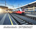 High-speed red passenger train at railway station platform under clear blue sky at sunset. Train station. Modern railway transportation concept. Railroad. Commercial. Urban rail transport in Austria