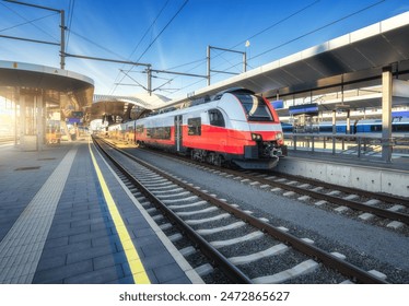 High-speed red passenger train at railway station platform under clear blue sky at sunset. Train station. Modern railway transportation concept. Railroad. Commercial. Urban rail transport in Austria