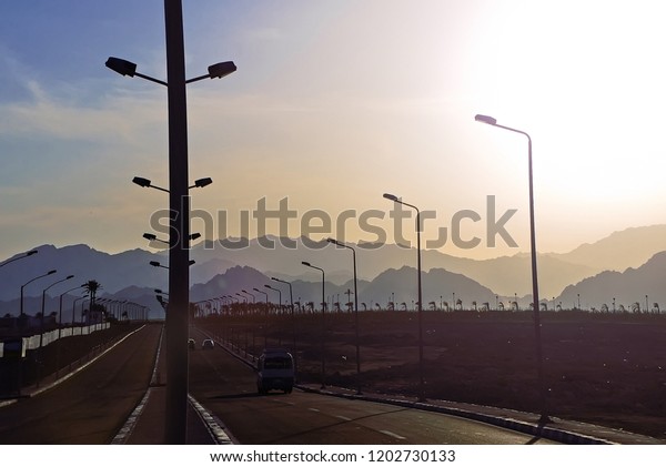 High-speed road highway with city\
lighting poles lamps. High mountains in the fog against the\
background, sunset, twilight, landscape, aerial perspective,\
cars