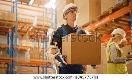 High-Tech Futuristic Warehouse: Worker Wearing Advanced Full Body Powered exoskeleton, Walks with Heavy Cardboard Box. Exosuit amplifies Human strength. Low Angle Portrait Shot