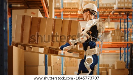 High-Tech Futuristic Warehouse: Worker Wearing Advanced Full Body Powered exoskeleton, Lifts Heavy Pallet full of Cardboard Boxes. Delivery Exosuit amplifies strength.
