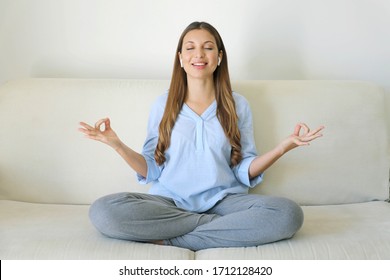 High-Tech Meditation. Young Woman With Wireless Earphones Using Meditation App On Smart Phone Sitting In Lotus Position On Confortable Sofa. Peaceful Millennial Girl Doing Yoga Exercises Alone.