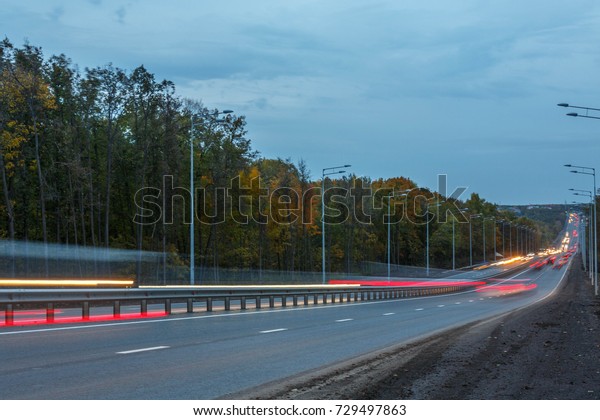 Highway in the evening in October,\
cars headlights on, lanterns and trees along the\
highway.