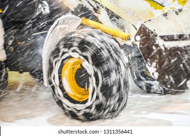 Highway maintenance heavy truck cleaning road during blizzard or snowstorm in evening, winter transport calamity removing in center of city. Big tire with chains is moving.