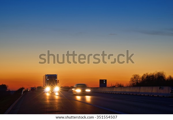 Highway with oncoming trucks and a car after\
sunset. Shining the spotlight cars. Information boards about the\
temperature. Blue and orange sky is\
clear.