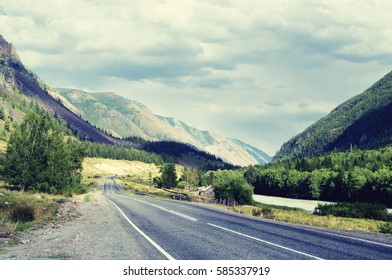 Highway road in a narrow mountain valley along the river, woodland and farm at sunset under a cloudy sky with blue clouds Chuyskiy trakt Altai, Siberia, Russia