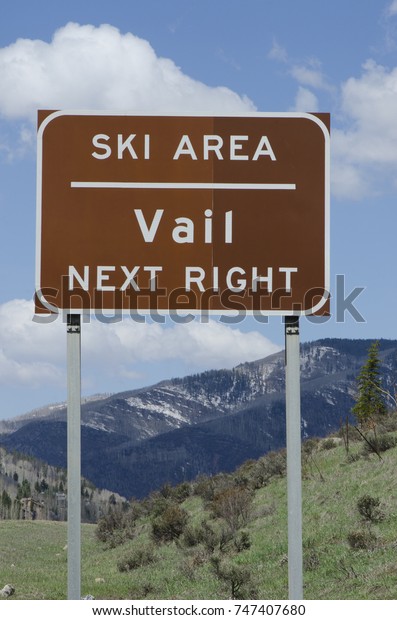 A highway sign announcing Vail Ski Area is\
photographed in the spring, with mountain scenery and billowing\
clouds beyond.
