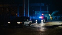 Highway Traffic Patrol Car In Pursuit Of Criminal Vehicle, Traffic Stop, Pull Over, Arrest. Police Officer Gets Out Of Squad Car, Approaches Suspect. Cinematic Action In Industrial Urban Area At Night