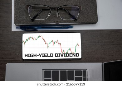 High-yield dividend concept. Top view of stocks price candlestick chart in phone on table near laptop, notepad and glasses with inscription high-yield dividend. Business, finance concept.