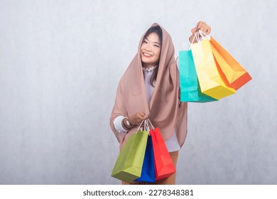 hijab girl smiling thinly lifting next paperbag shopping. portrait Asian Chinese Muslim Muslim hijab headscarf woman fasting Ramadan on Eid Al-Fitr day with white background.
