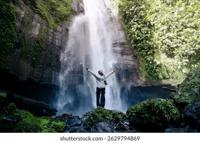 Una mujer con hiyab vestida de verde salvia se para con los brazos bien abiertos debajo de una cascada alta y poderosa. Captada desde atrás, la Imagen resalta la armonía entre lo humano y la naturaleza