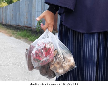 A hijab-wearing woman, with a light step, returned from the market carrying several transparent plastic bags filled with her purchases. Inside, there were purple sweet potatoes, fresh red chilies, - Powered by Shutterstock