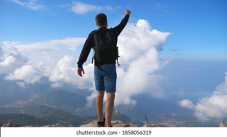 Hiker With Backpack Reaching Up Edge Of Mountain And Victoriously Raising His Hand. Man Actively Resting During Travel Enjoying Freedom On Hill Top. Scenic Landscape At Background. Slow Mo Back View